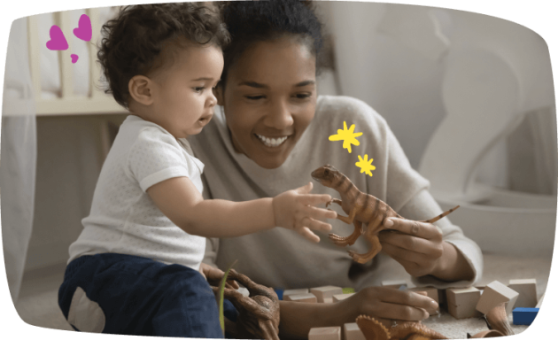 Uma babá sorridente segura um dinossauro de brinquedo para um bebê curioso, sentados juntos no chão rodeados de blocos de madeira e outros dinossauros.